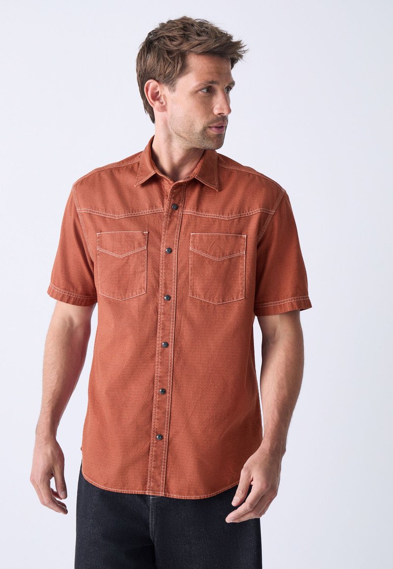Short-sleeve, rust-colored shirt in textured fabric, featuring two chest pockets, black buttons, and contrasting stitching. Casual fit.