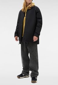 Zwarte donsjas met capuchon, gedragen over een gele hoodie. Gecombineerd met loszittende grijze jeans en zwarte sneakers met witte accenten.