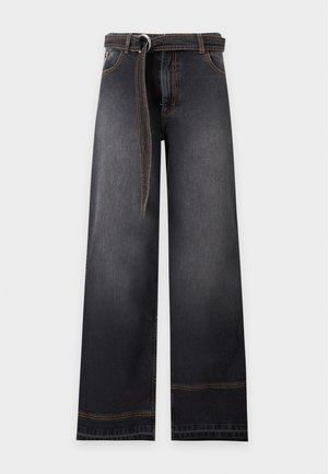 Femme aux cheveux courts portant un haut noir sans manches, un jean large foncé avec une ceinture et des chaussures rouge foncé, debout les mains dans les poches.