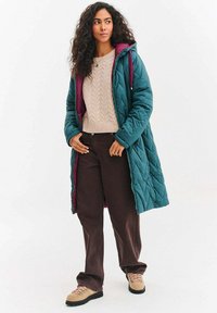 Dlouhý tyrkysový prošívaný kabát s bordovou podšívkou, kombinovaný s béžovým svetr s copánkovým vzorem, hnědé široké kalhoty a béžové turistické boty.