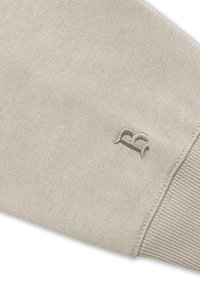 Beige Baumwoll-Sweatshirtärmel mit geripptem Bündchen, das ein dezentes, gesticktes Logo in derselben Farbe präsentiert. Glatte Textur und schlichtes Design.