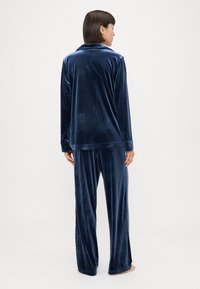 Conjunto de pijama em veludo azul-marinho, com mangas compridas, blusa com colarinho e calças largas, com uma textura suave e macia.