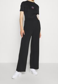 Černé široké kalhoty s elastickým pasem, doplněné o přiléhavé černé tričko s vícebarevným logem. Bílí tenisky dokončují celý vzhled.