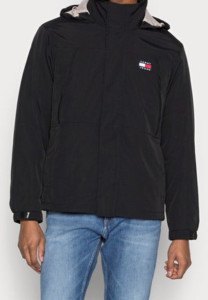 Mann trägt schwarze Kapuzenjacke mit Tommy Jeans-Logo und blaue Jeans, Hände entspannt an den Seiten, vor schlichter Hintergrung.