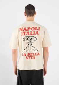 Beige bomull T-skjorte med fet rød tekst "NAPOLI ITALIA" og "LA BELLA VITA" samt et grafisk bilde av en pizzafformet vulkan på baksiden.