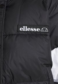 Doudoune noire avec écusson logo "ellesse" brodé en blanc sur la poitrine, montrant une partie des coutures avant et des manches.