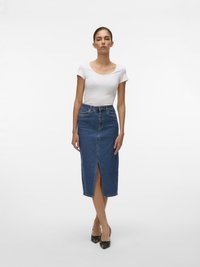 Femeie stând cu fața înainte, purtând un top alb cu mâneci scurte, croială strâmtă, o fustă midi din denim albastru cu talie înaltă și slit frontal, și pantofi cu toc negri.