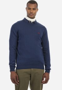 Mörkblått stickat tröja med rundad halsringning, åtsittande ärmar och ribbat nederkant. Har en liten röd logotyp på vänster bröst.