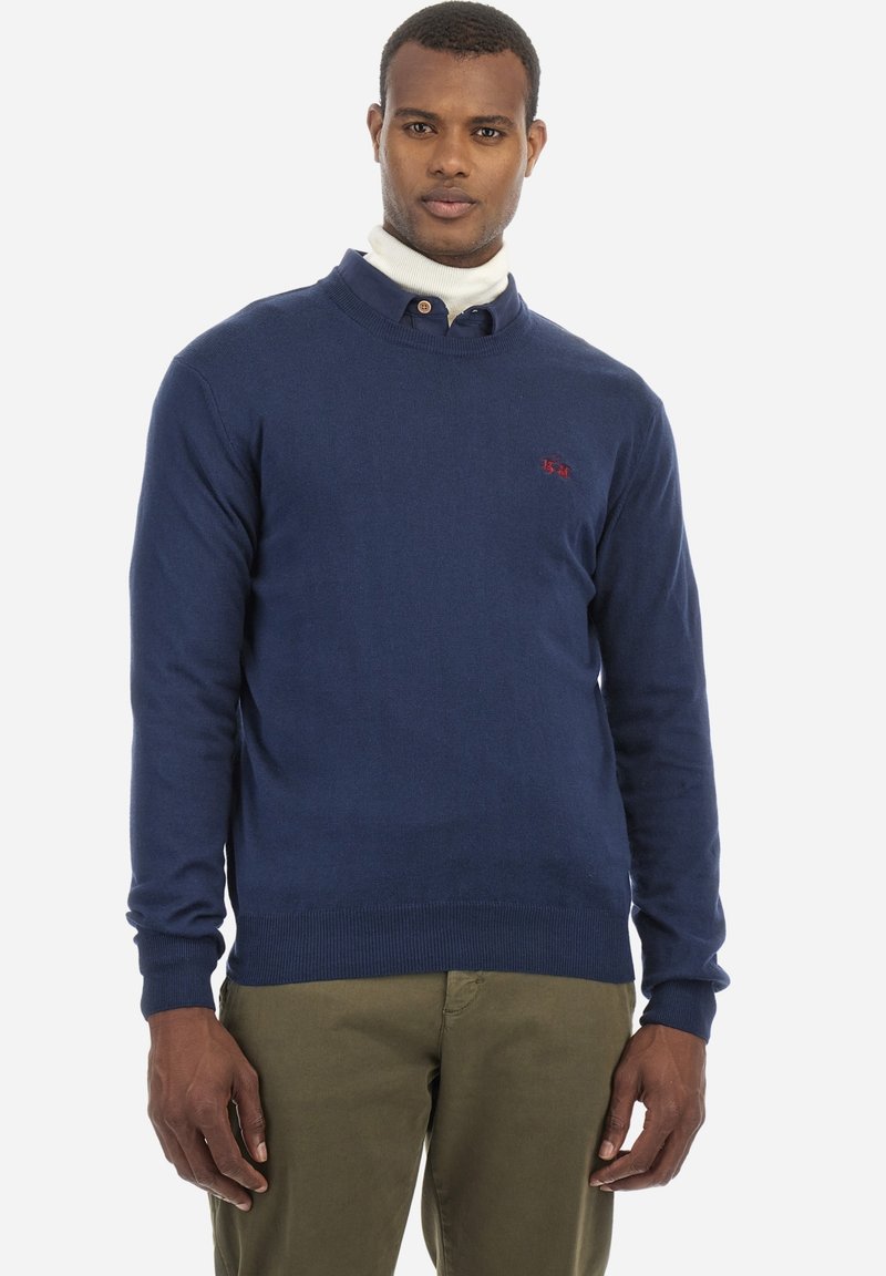 Mörkblått stickat tröja med rundad halsringning, åtsittande ärmar och ribbat nederkant. Har en liten röd logotyp på vänster bröst.