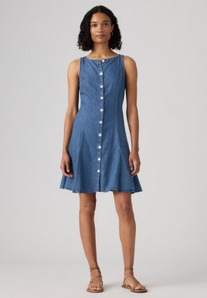 Mujer de pie vestida con un vestido vaquero azul sin mangas con botones delanteros y sandalias planas marrones con tiras sobre un fondo claro y liso.