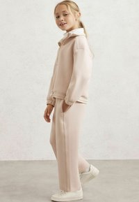 Šviesiai rožinė treniruočių komplektas, susidedantis iš užtrauktuku užsegamo džemperio su gobtuvu ir derančių sportinių kelnių su šoninėmis kišenėmis bei elastingomis apykaklėmis. Papildyta baltais sportbačiais.