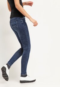 Jeans ajustados en denim oscuro con un ligero desgaste. Presentan un diseño estándar de cinco bolsillos y un corte slim, combinados con zapatillas blancas y calcetines con estampado.
