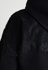 Črna kapuca z zapletenim črnim čipkastim detajlom na ramenskih delih, ki združuje mehkobo blaga z teksturiranim čipkastim kontrastom.