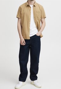 Camicia button-up a maniche corte color beige con un tessuto testurizzato, abbinata a jeans blu scuro e scarpe da ginnastica bianche con dettagli gialli.
