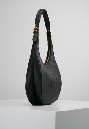 Sac à bandoulière en cuir noir avec sangle réglable, boucles dorées et logo embossé, posé sur un piédestal blanc sur fond gris.