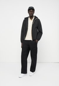 Chaqueta acolchada negra con capucha, camiseta polo beige claro, pantalones cargo negros y zapatillas blancas. Una gorra simple completa el atuendo.