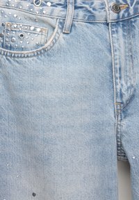 Jean en denim bleu clair avec des décorations clous argentés près de la poche avant et une fermeture par bouton en métal.