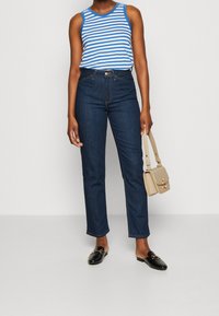 Top senza maniche a righe blu e bianche, jeans scuri a vita alta, mocassini neri e una borsa a tracolla beige, in piedi su uno sfondo chiaro.