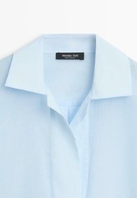 Chemise bleu clair à col avec patte de boutonnage cachée et étiquette noire indiquant Massimo Dutti fabriqué au Maroc.