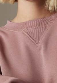 Close-up van een persoon die een mauve sweater draagt met een ronde hals en geribbelde V-detail onder de kraag, met kort blond haar zichtbaar.