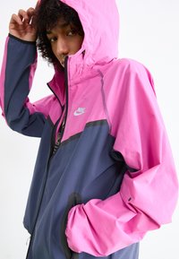 Nike jakke med et design i pink og marineblå farveblok, lavet af letvægtsmateriale med hætte, fuld lynlås og elastiske manchetter.