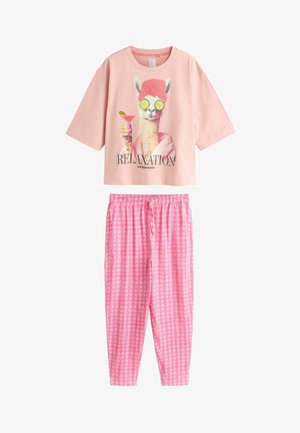 Frau mit kurzen dunklen Haaren, die ein pinkes T-Shirt mit Lama-Motiv, rosa karierte Hosen und passende pinke Hausschuhe trägt, steht vor einer hellen Wand.