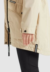 Beżowa, wodoodporna kurtka zapinana na zamek i napy, z regulowanym ściągaczem, czarnymi akcentami i widoczną metką marki.