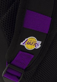 Sac à dos en mesh noir avec une sangle violette et un logo des Los Angeles Lakers, accompagné d'un graphisme de basket-ball, mettant en avant l'identité de l'équipe et ses couleurs.
