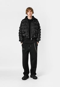 Chaqueta bomber negra con tiras de tela en capas, cierre de cremallera; usada sobre un suéter negro y pantalones a juego con bolsillos laterales y detalle de cadena.