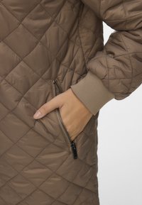 Handen i den zippered fickan på en brun quiltad jacka med ribbad manschett på ärmen.