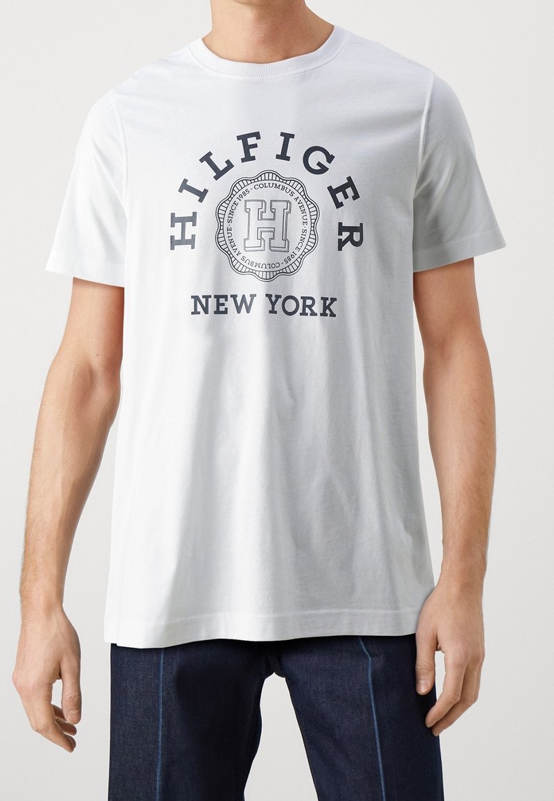 White cotton T-shirt featuring a circular graphic design with “HILFIGER” and “NEW YORK” in navy text. Short sleeves and round neckline.