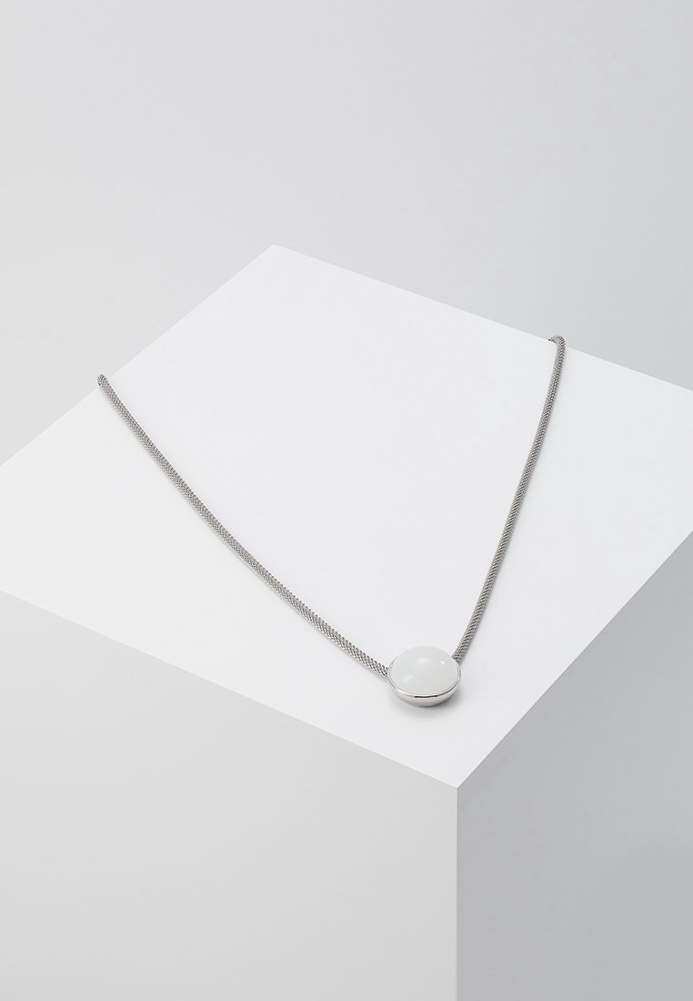 Collier en chaîne argentée avec un pendentif blanc lisse et rond reposant sur un piédestal cubique blanc sur fond gris clair.