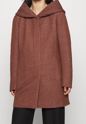 Femme portant un manteau en laine marron mi-long avec une grande capuche et des manches longues, associé à un pantalon noir.