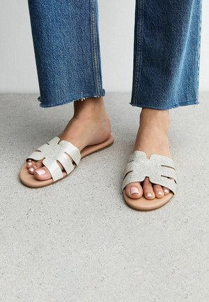 Glitrende beige slidesandaler har et udskæringsdesign og en flad, tan sål. Kombinationen er med slidte blå jeans på en tekstureret overflade.