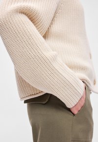 Μπεζ πλεκτό κιμονό με υφαντό σχέδιο, διαθέτοντας χαλαρή εφαρμογή και στρογγυλή άκρη, συνδυασμένο με ελαιόχρωμα παντελόνια με πλαϊνές τσέπες.