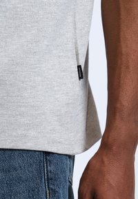 Camiseta gris claro hecha de una suave mezcla de algodón, que presenta una abertura lateral y una pequeña etiqueta de la marca negra cerca del dobladillo. Combina con denim.