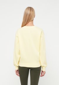 Geltonas megztinis su ilgomis rankovėmis, sutrauktomis riešais ir apačia, bei apvalia iškirpte, derinamas su tamsiai žaliais prigludusiais kelnių.