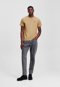 Korte mouwen tan T-shirt met een klein logo op de borst, gecombineerd met grijze jeans en zwart suède schoenen. Eenvoudige, casual pasvorm.