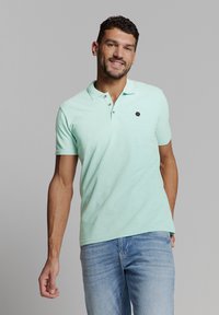 Mintgroene poloshirt met een drieknopenplacket, geweven stof en een klein zwart embleem op de borst. Draag dit met lichtblauwe jeans.