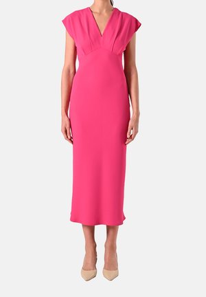Frau trägt ein leuchtend pinkes Midikleid mit kurzen Kappenärmeln, einem tiefen V-Ausschnitt und beigen spitzen Pumps und steht vor einem schlichten Hintergrund.