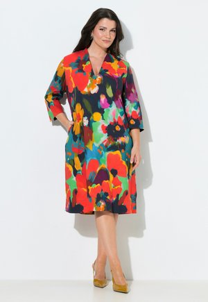 Kobieta stojąca z jedną ręką w kieszeni, ubrana w kolorową, kwiatową sukienkę do kolan i musztardowo-żółte szpilki na białym tle.