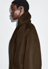 Manteau en laine marron avec un col montant et replié. Le design est oversize, avec une texture lisse et sans fermetures ni embellissements visibles.
