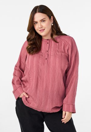 Kvinde iført en struktureret langærmet bluse i rose pink med knapper, sorte bukser og sølvøreringe, der poserer med den ene hånd i lommen.