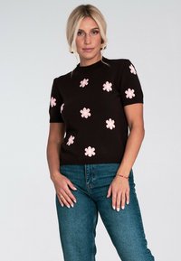 Kobieta w czarnej krótkiej bluzie z różowymi kwiatowymi wzorami i niebieskich dżinsach, stojąca na tle jasnego, jednolitego tła.