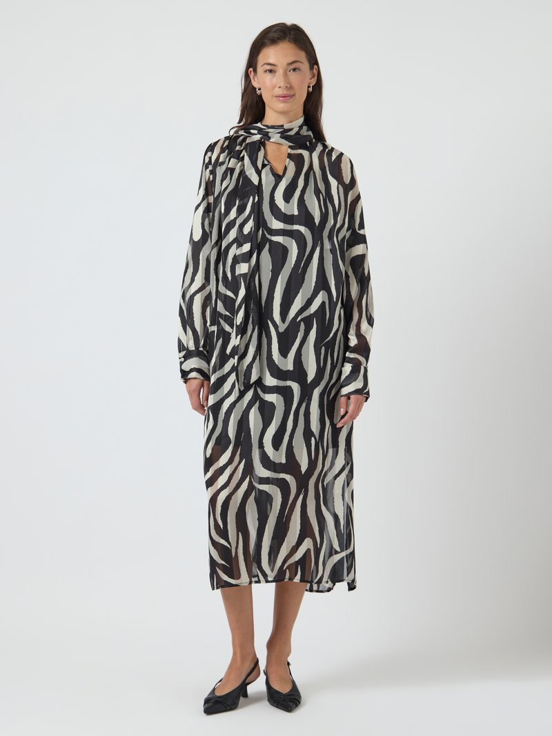 Vestido maxi com estampado de zebra preto e branco, com gola de laço e mangas compridas, feito de tecido leve e transparente. Sapatos pretos de salto bloco.