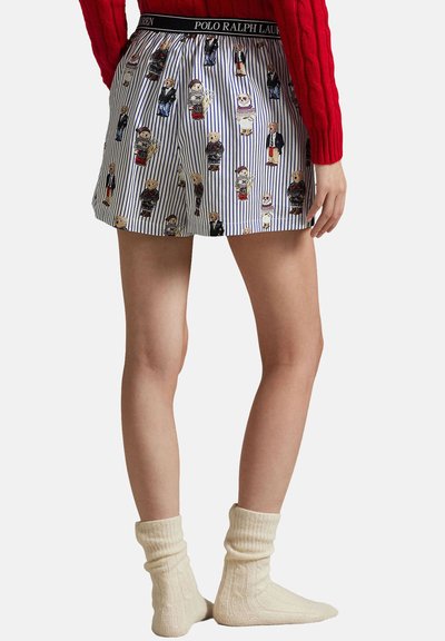 Person som bär randiga shorts med björnillustrationer, en röd tröja och cremefärgade stickade strumpor, stående mot en enfärgad bakgrund.