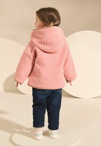 Rosa Fleece-Hoodie mit texturiertem Stoff, kombiniert mit dunkelblauen Jeans und weißen Sneakers. Das Outfit hat einen lässigen Schnitt und umgekrempelte Ärmel.