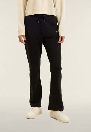 Persona che indossa pantaloni neri con coulisse, maglia a maniche lunghe color crema e sneakers bianche, in piedi davanti a uno sfondo semplice.