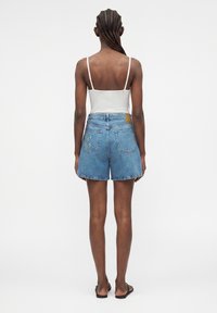 Femme aux cheveux tressés portant un débardeur blanc à fines bretelles, un short en jean bleu clair brodé et des tongs noires, debout de dos.