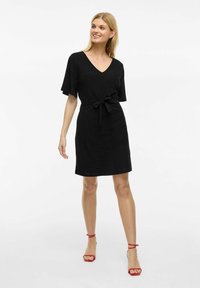 Svart kortärmad klänning med V-ringning och en knytrem vid midjan. Slät textur med måttlig längd, parat med röda ankelbandsskor.